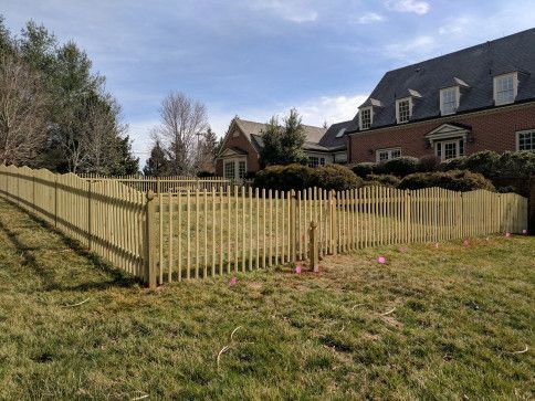 Wooden picket fence enclosing a grassy yard, with a large brick house in the background.