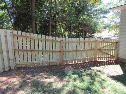 Wooden picket fence with gate enclosing a grassy yard, backed by trees, in daylight.