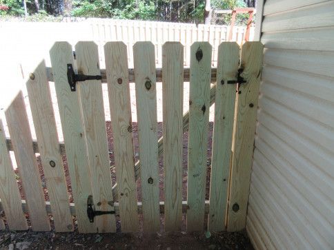 Wooden picket gate with black hinges, attached to a siding-covered wall.