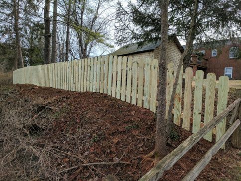 Wooden picket fence bordering a brown dirt hill, with trees and a brick building in the background.