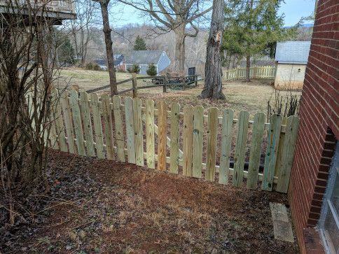 Wooden picket fence next to a brick building. Brown grass and trees in the background.