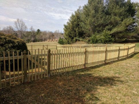 Wooden picket fence surrounding a grassy yard with trees in the background under a blue sky.
