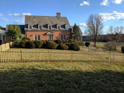 Red brick house with a picket fence, bushes, and a bare tree under a blue sky.