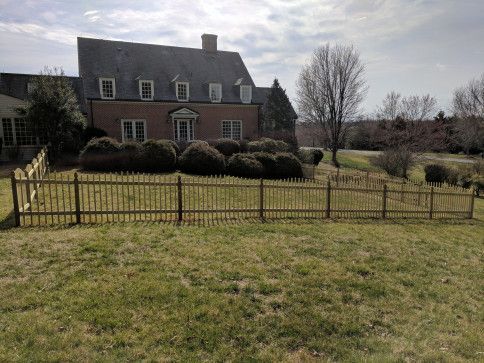 A two-story brick house with a wooden fence in front, set on a grassy lawn under a partly cloudy sky.