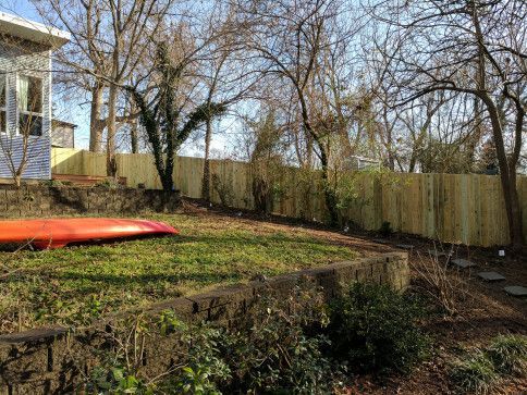 A backyard with a wooden fence, grass, and trees on a sunny day. An orange kayak rests in the yard.