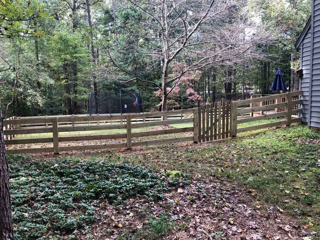 Wooden fence surrounds a yard with a gate, trees in the background, and a house to the right.