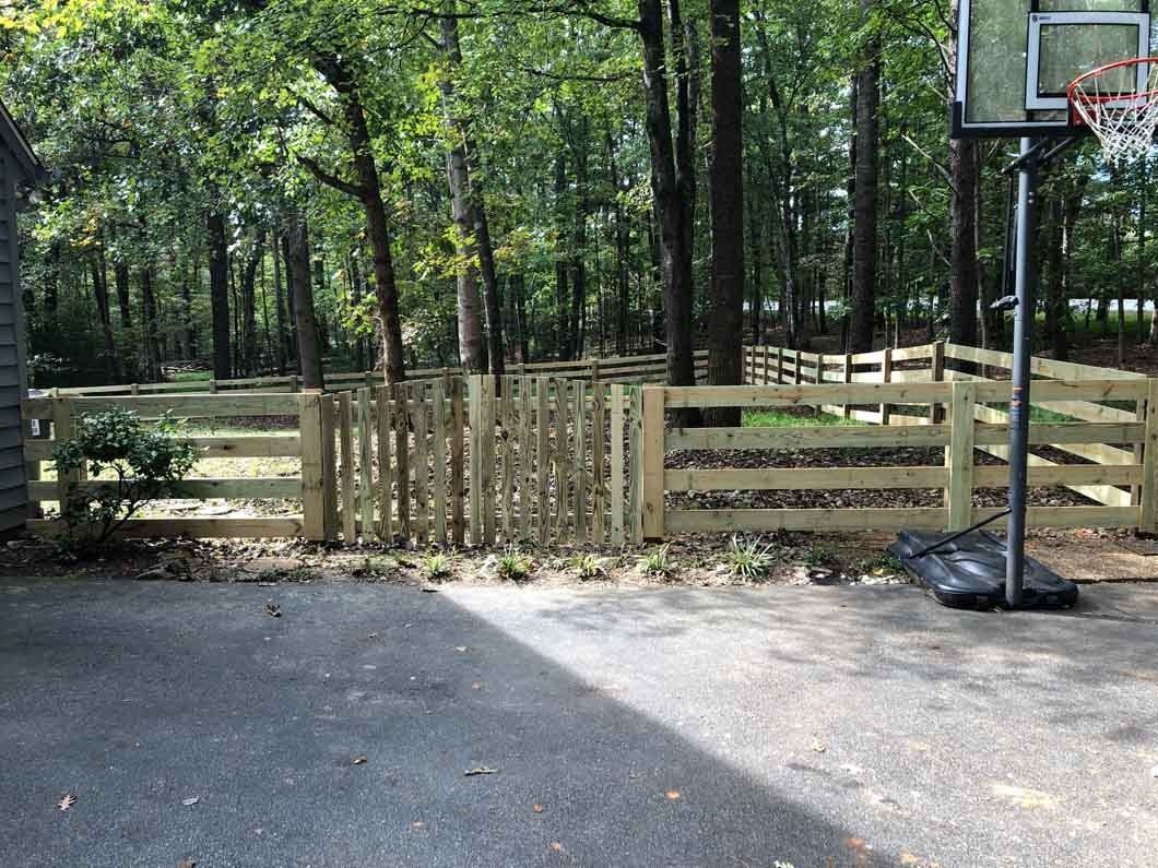 Wooden fence with gate, basketball hoop, and driveway leading into a wooded area.