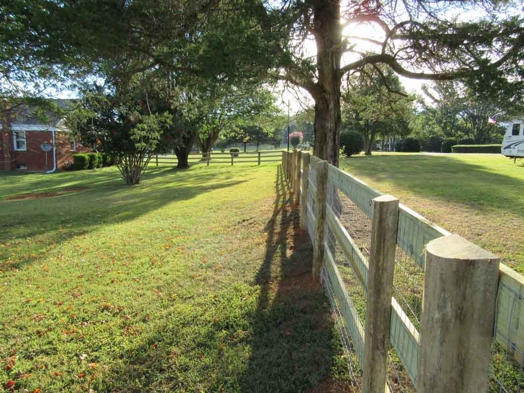 Wooden fence in a grassy field with trees and a building in the background on a sunny day.
