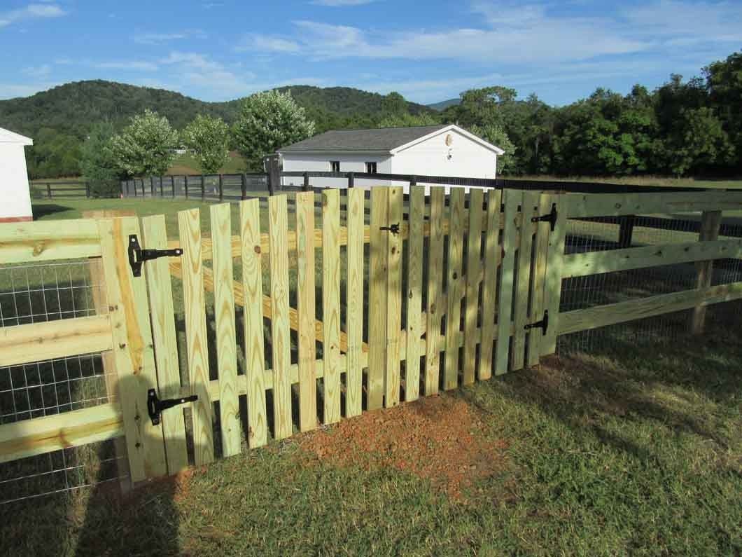Wooden gate in a grassy field, attached to a fence with a white building and hills in the background.
