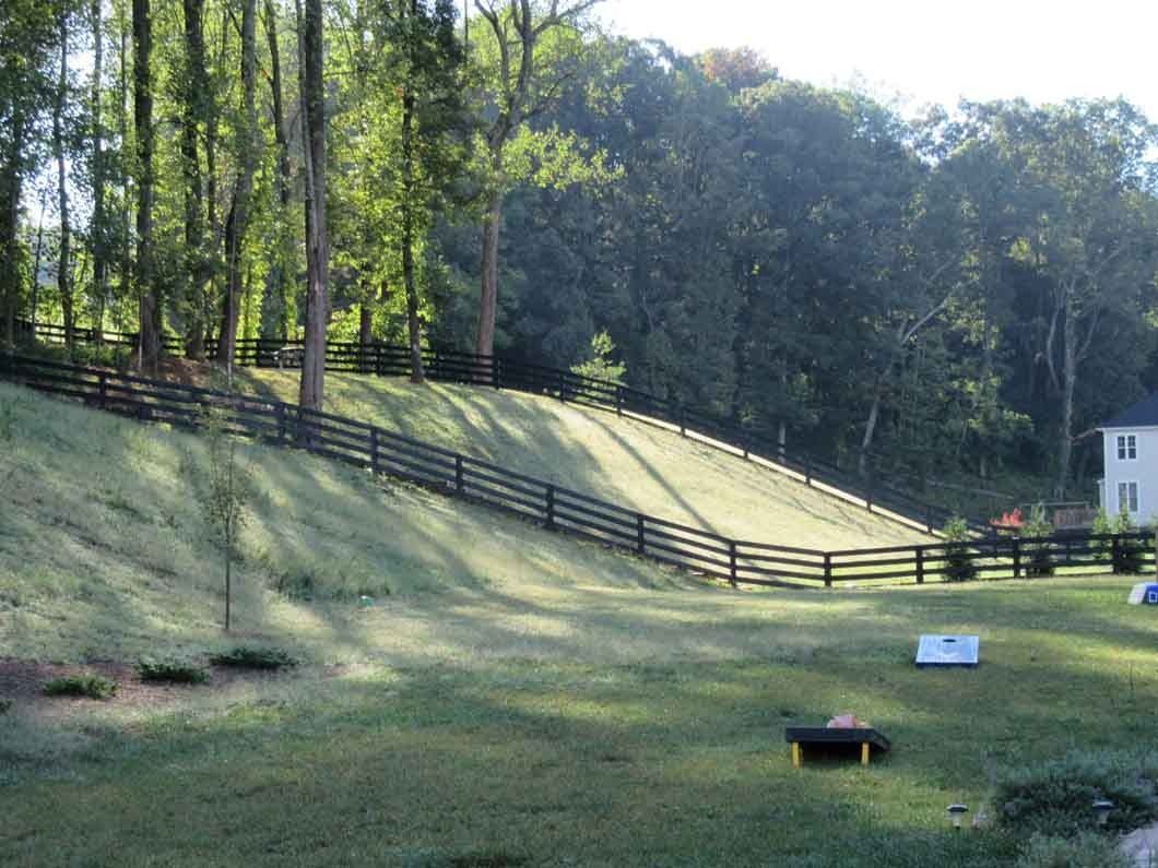 Grassy hill with black fence and trees, sunlight. A house is visible in the background.