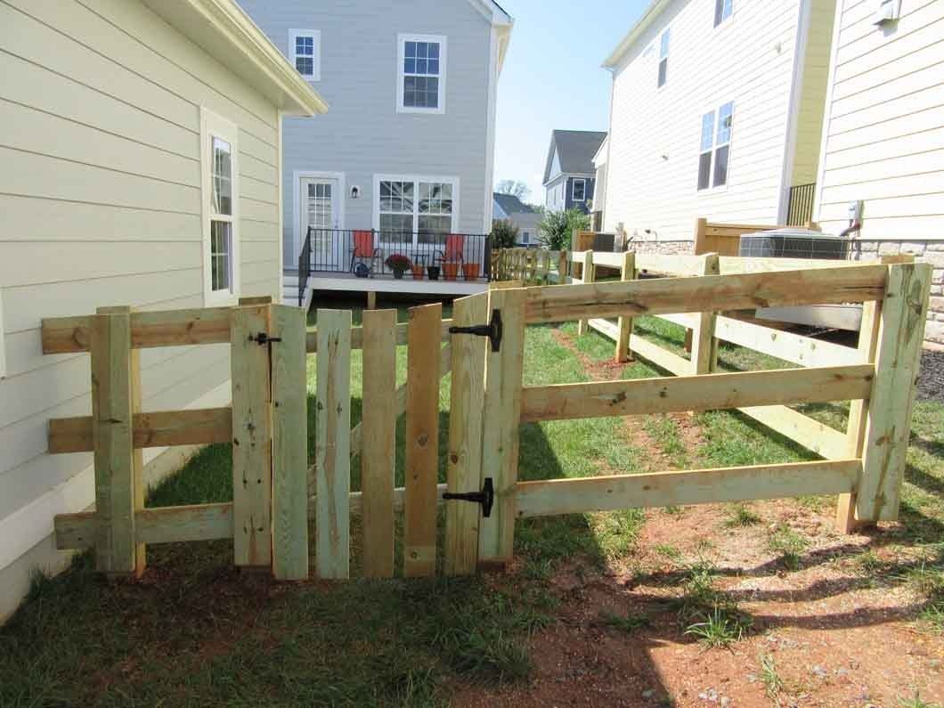 Wooden fence with gate in a yard, connecting to a house; grass and other houses in background.