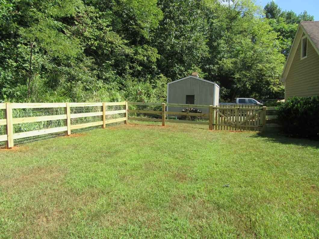 Wooden fence encloses a grassy yard with a shed and trees in the background. A house is on the right.