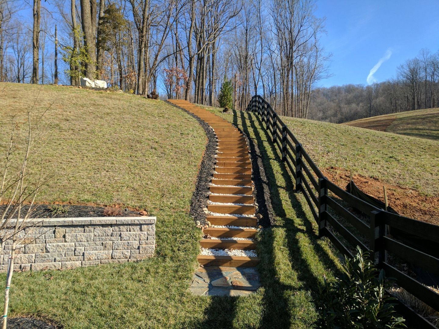 Wooden staircase ascending a grassy hill, with a black fence alongside and trees in the background.