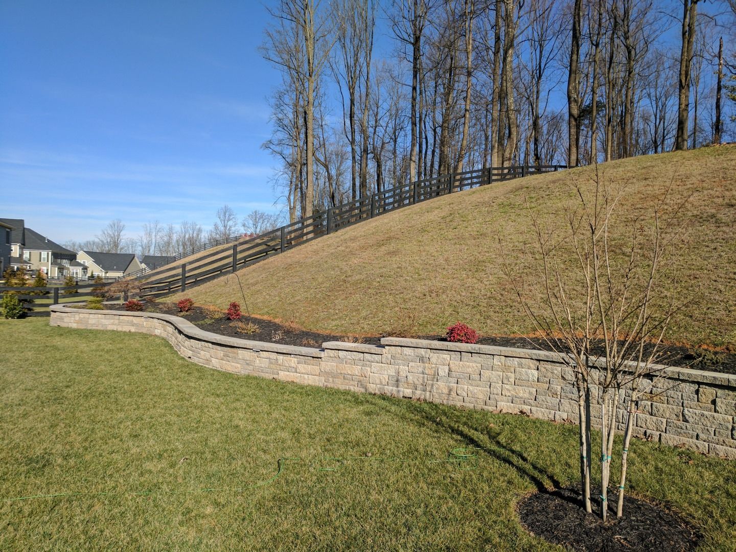 A landscaped backyard with a retaining wall, a hill with a fence, and a clear blue sky.