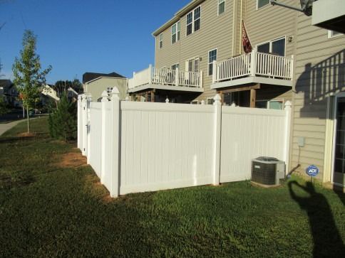 White vinyl fence encloses a backyard with air conditioning unit. Two-story house in background.