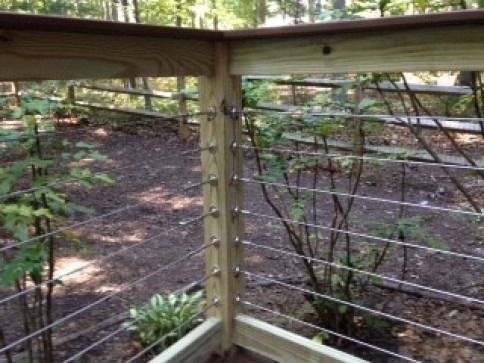 Wooden deck corner with stainless steel cable railing in a wooded setting.