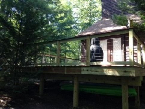 Wooden deck attached to a cabin, surrounded by trees. Two green kayaks sit below the deck.