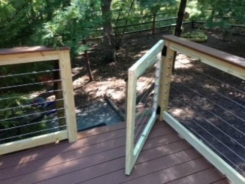Wooden deck with open gate, cable railing, and wooded background.