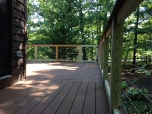 Wooden deck with cable railing overlooking trees. Brown deck boards, green foliage, bright sunlight.