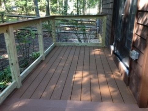 Wooden deck with railing, brown floorboards, next to a building. Forest background.