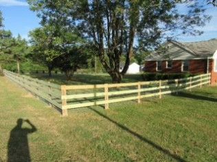 Green wooden fence surrounding a grassy yard with a house and trees in the background. A shadow is cast on the grass.