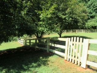 White fence with gate, enclosing a grassy area with trees, sunny outdoor setting.