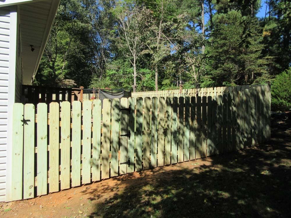 Wooden fence in a backyard, beside a house with a green lawn and trees in the background.