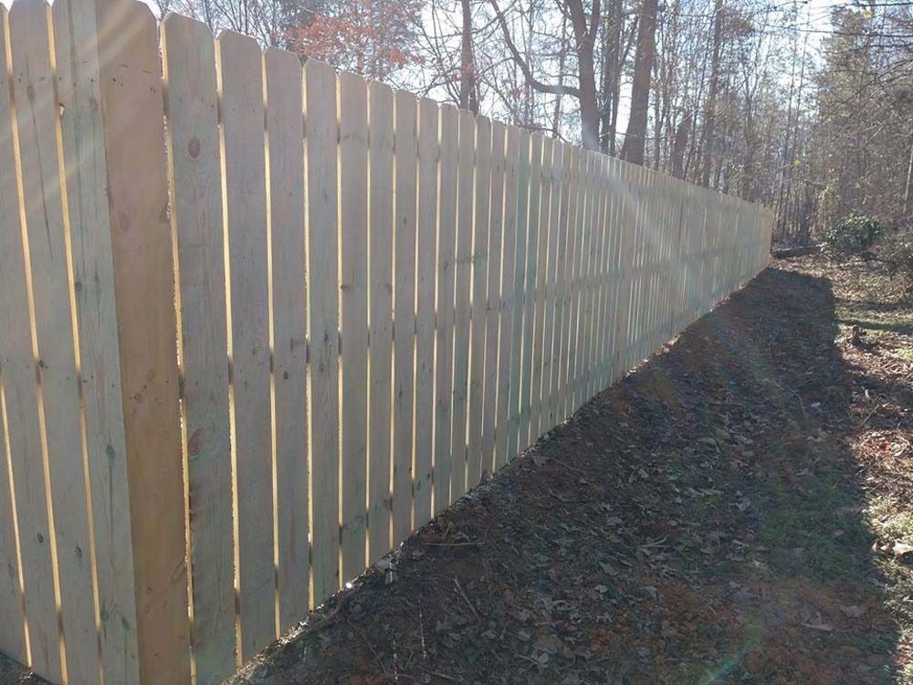 Wooden picket fence stretches along a wooded area, in the sunlight.