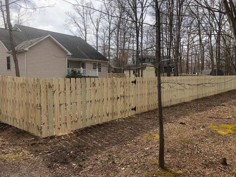 A wooden fence surrounds a tan house in a wooded area on a cloudy day.