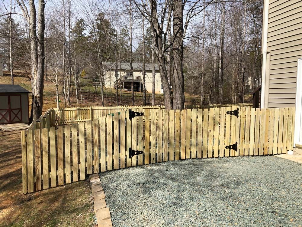 Wooden fence encloses a gravel patio area next to a house, with a wooded background.