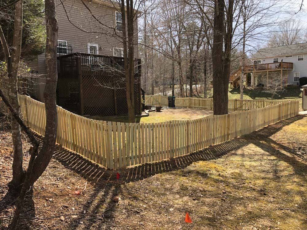 Wooden picket fence surrounds a backyard near a deck and trees.