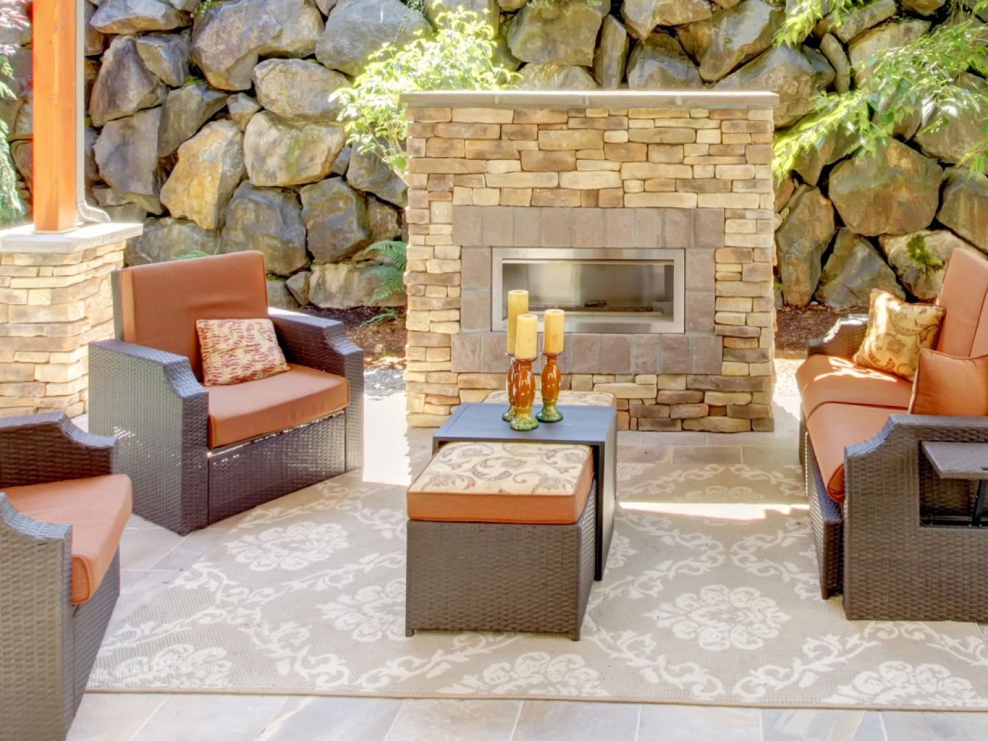 Outdoor patio with stone fireplace, dark wicker furniture with orange cushions, and patterned rug.