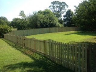 Wooden picket fence curving across a grassy yard, trees in the background under a blue sky.