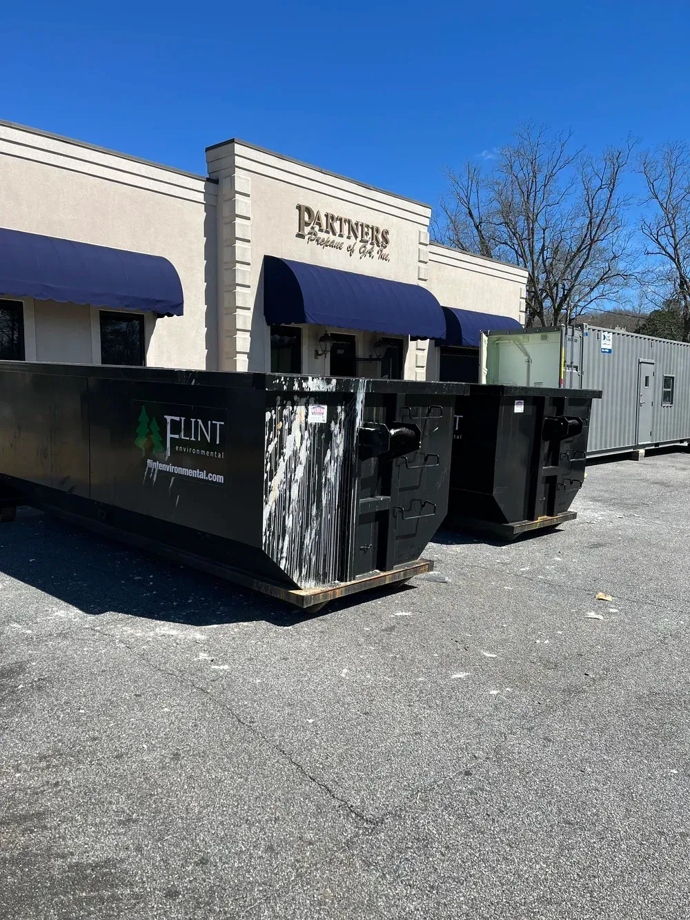 Two black dumpsters in front of a tan building with blue awnings and 