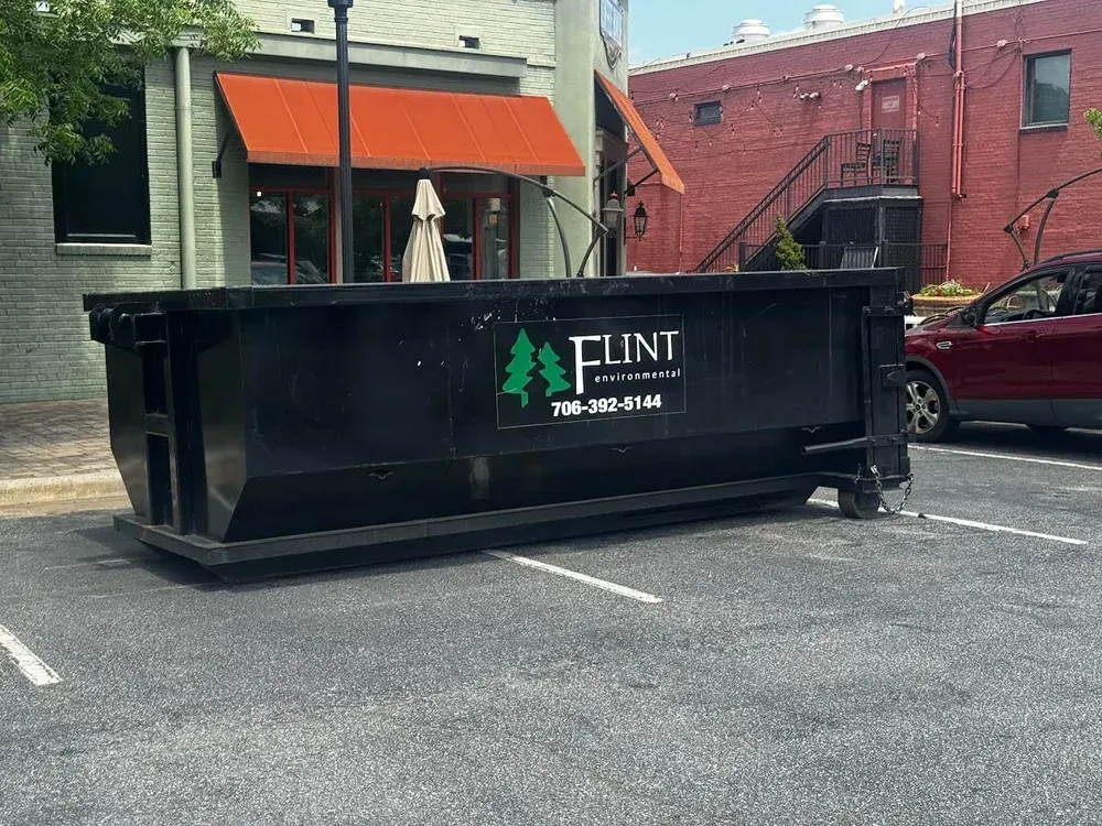 Black dumpster in parking lot, with business logo, next to building with red awning and car.