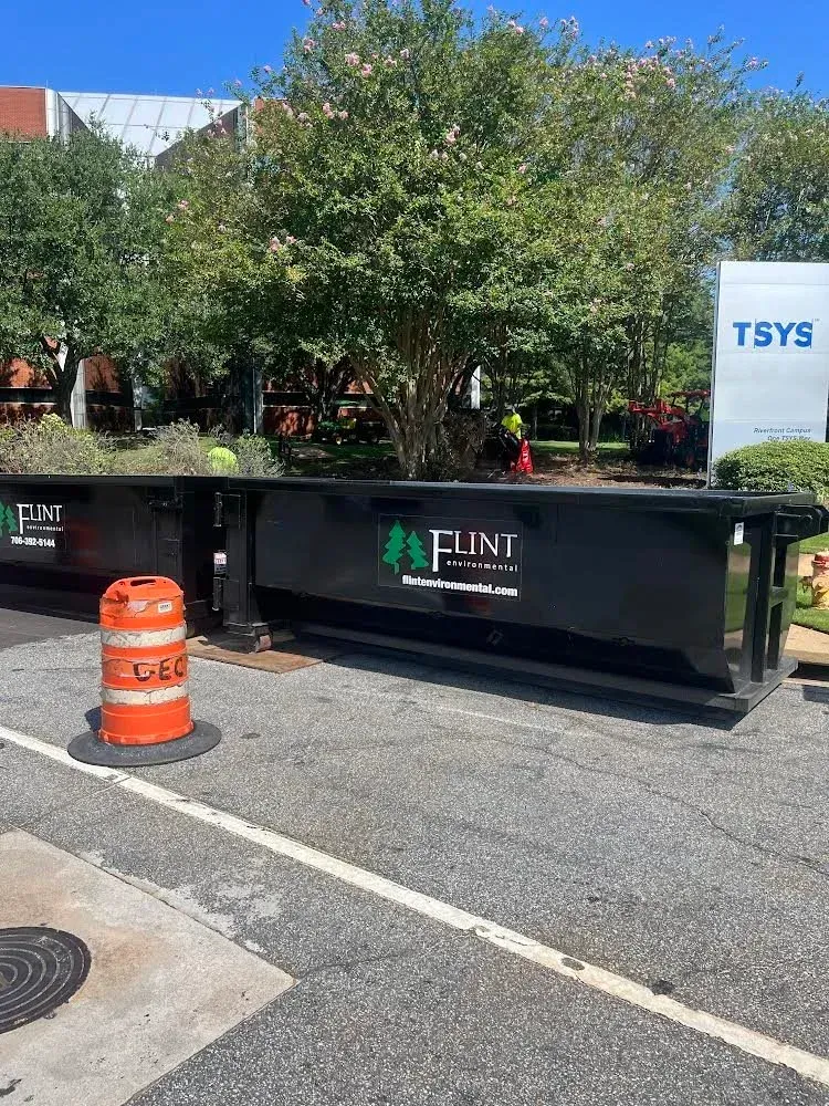 Two black dumpsters with Flint Landscaping logo, parked on asphalt with orange traffic cone; TSYS building in background.