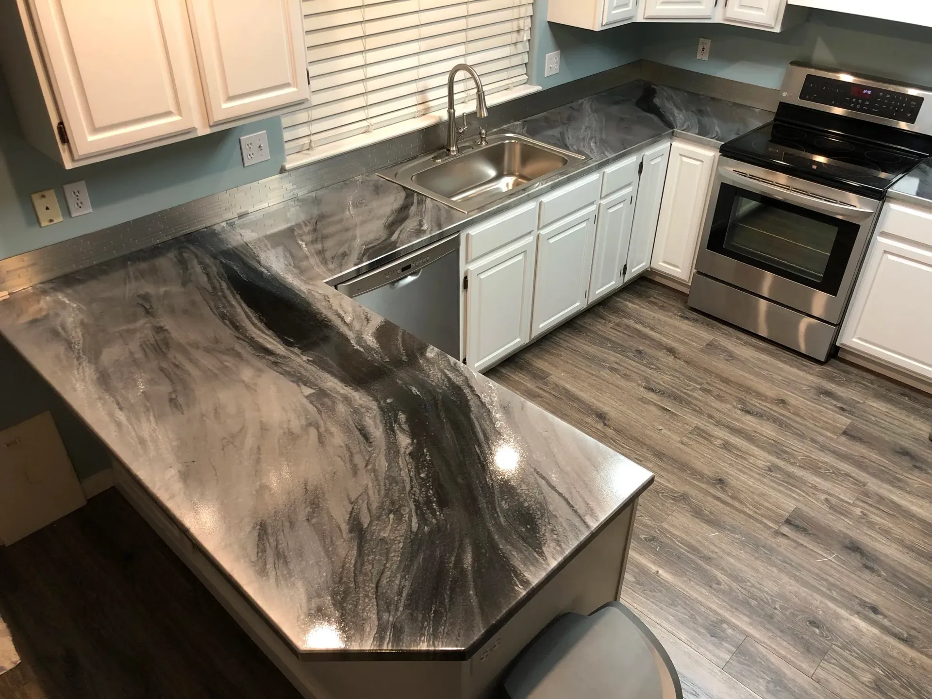 Modern kitchen with gray and silver countertop, stainless steel appliances, white cabinets, and wood-look flooring.