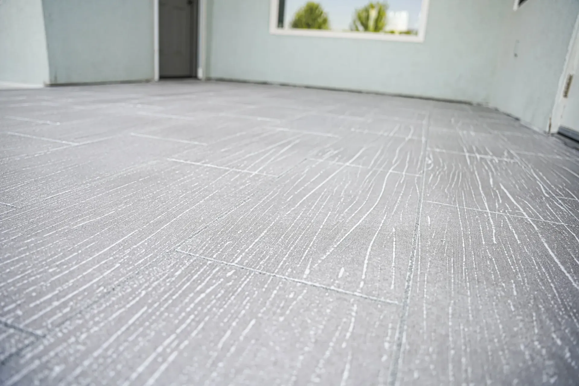 Gray flooring with scratch marks, in a room with light blue walls and a window.