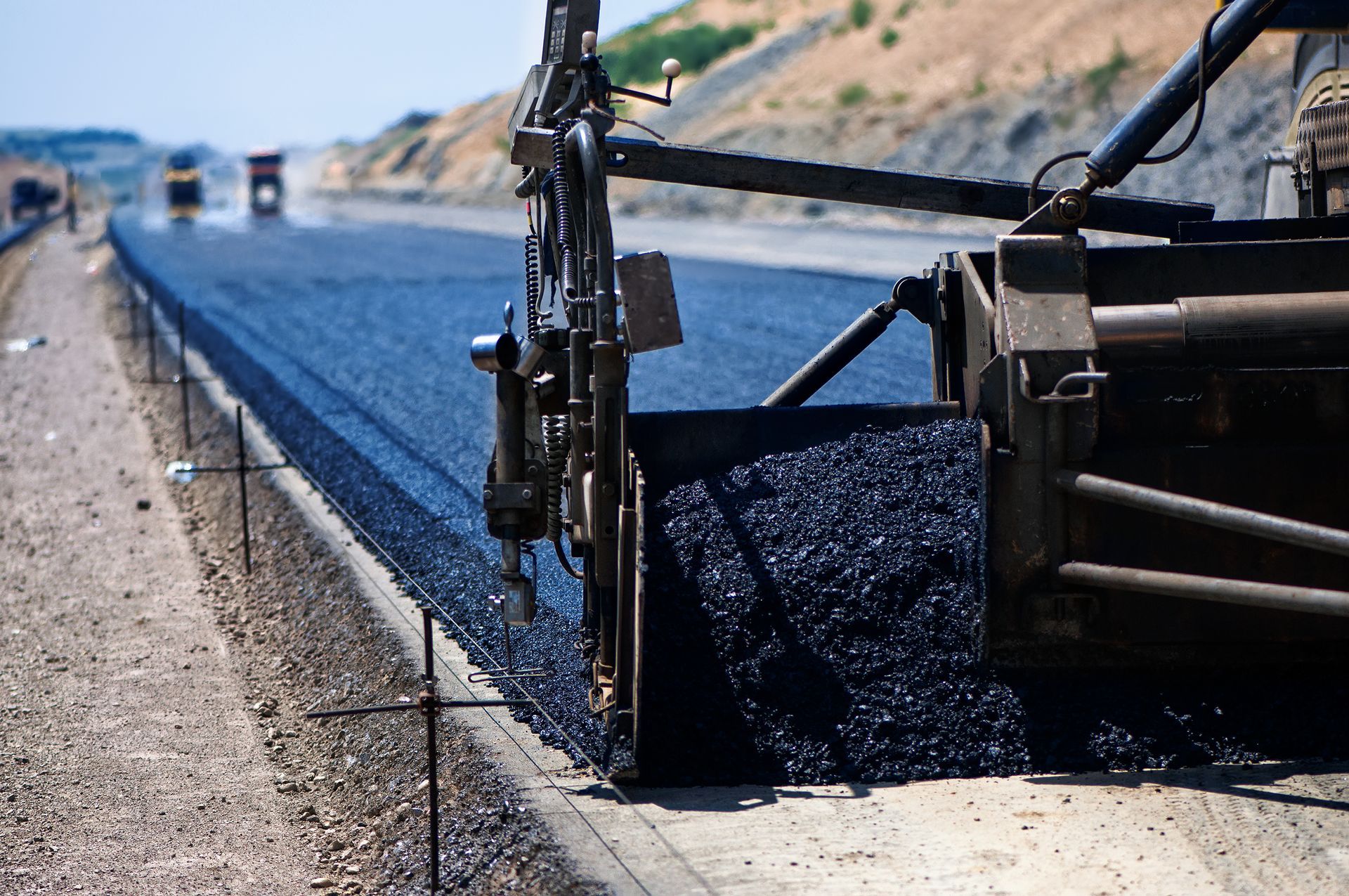 Road paving machine laying fresh asphalt on a highway with trucks in the background.