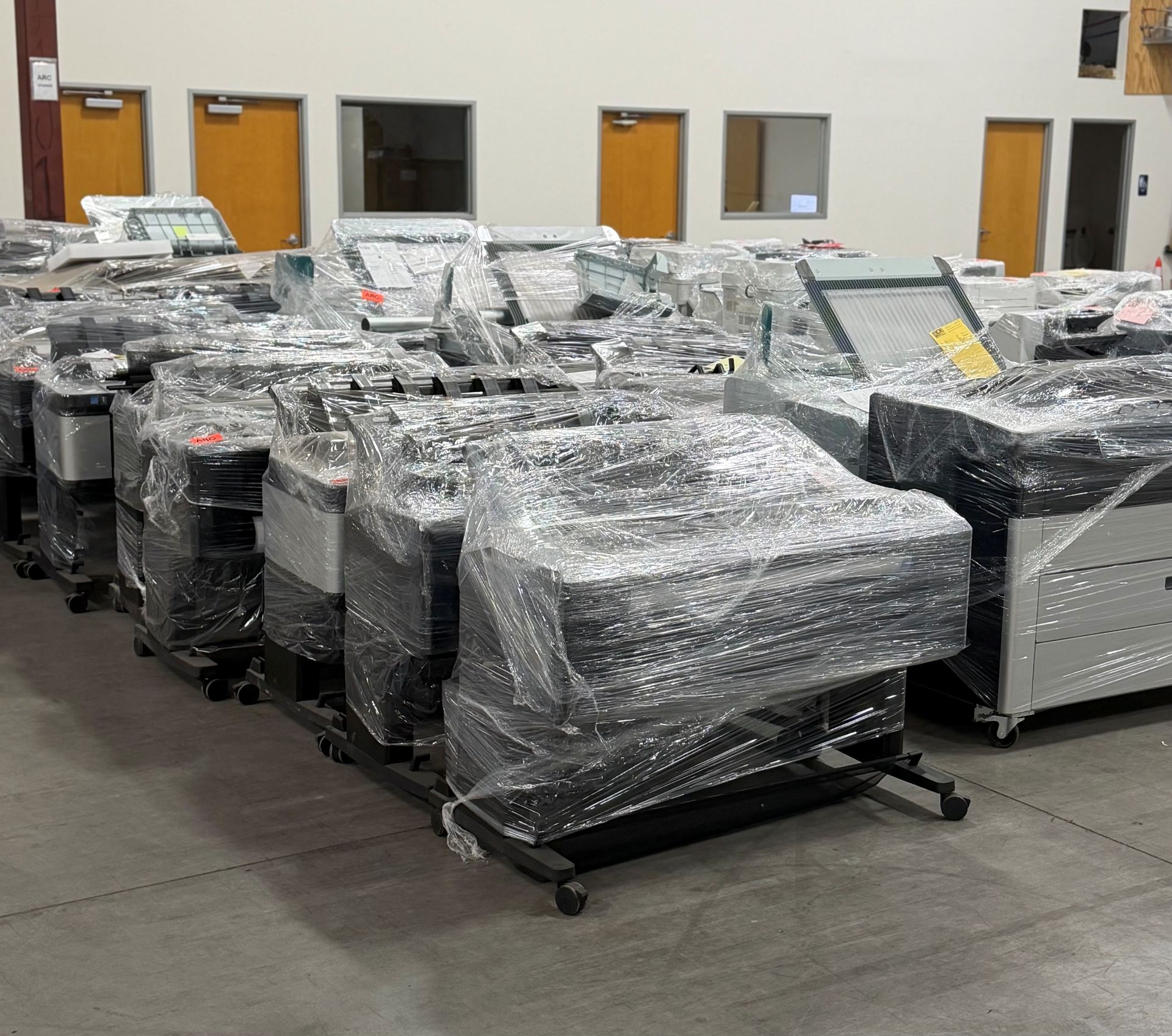 Wrapped industrial machines on a warehouse floor, lined up in rows near orange doors and windows.
