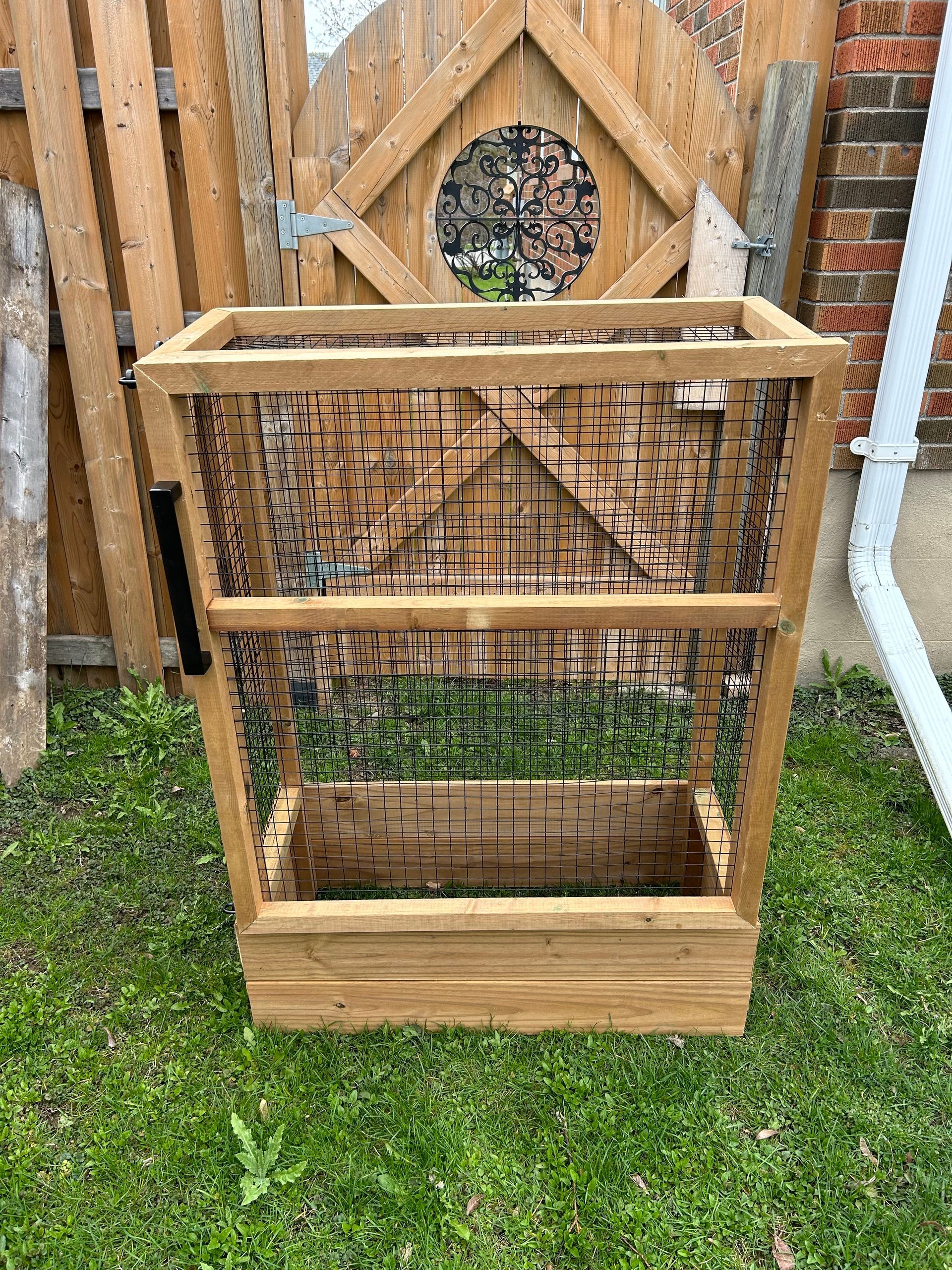 A wooden cage is sitting in the grass in front of a wooden fence.