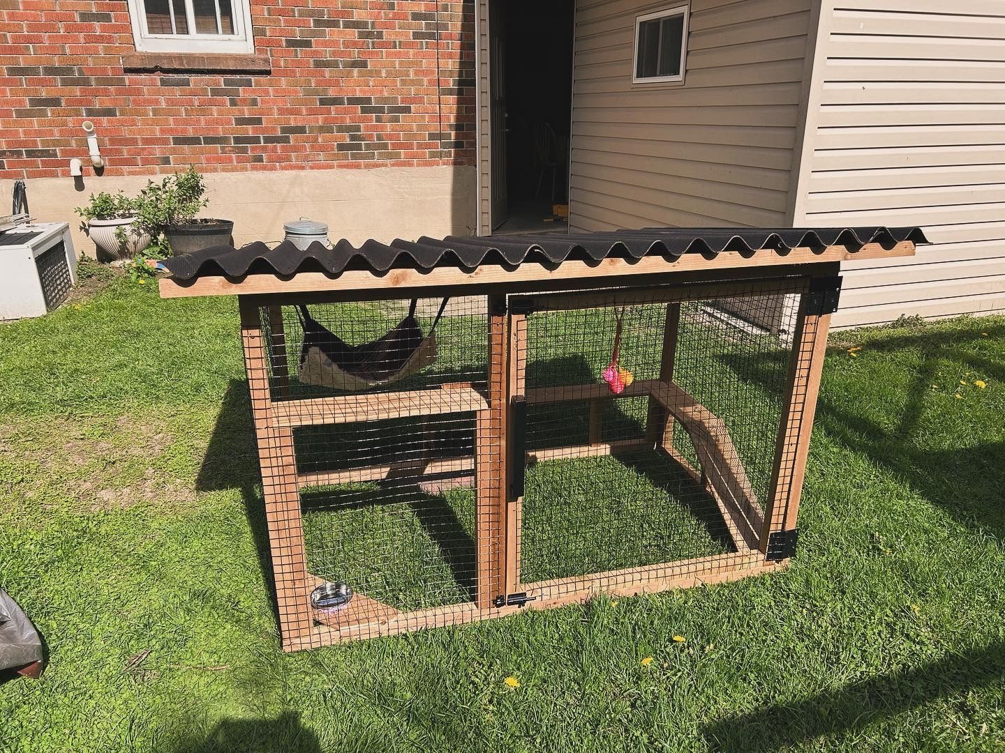 A wooden chicken coop is sitting in the grass in front of a brick building.