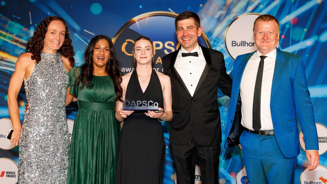 Five people in formalwear posing at an awards event, holding a trophy on a blue-lit stage.