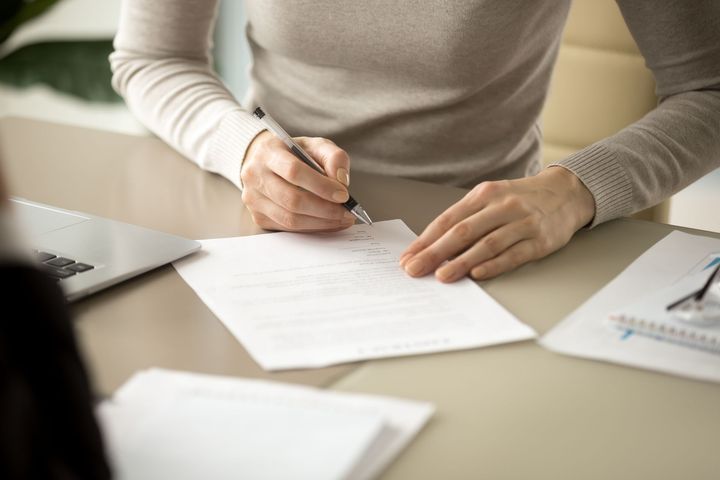 Person signing a document at a desk, with a laptop and papers.