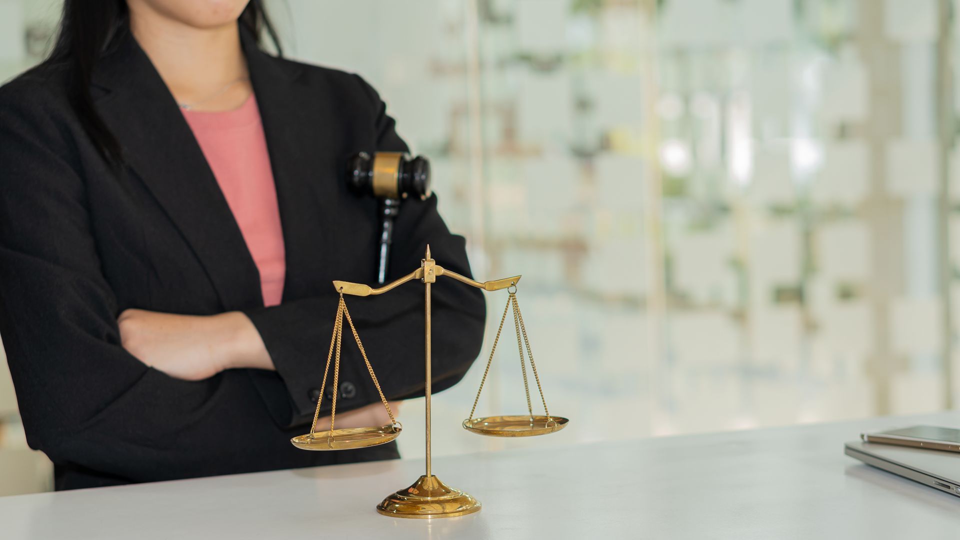 Lawyer in black blazer with arms crossed, next to scales of justice and gavel on a desk.