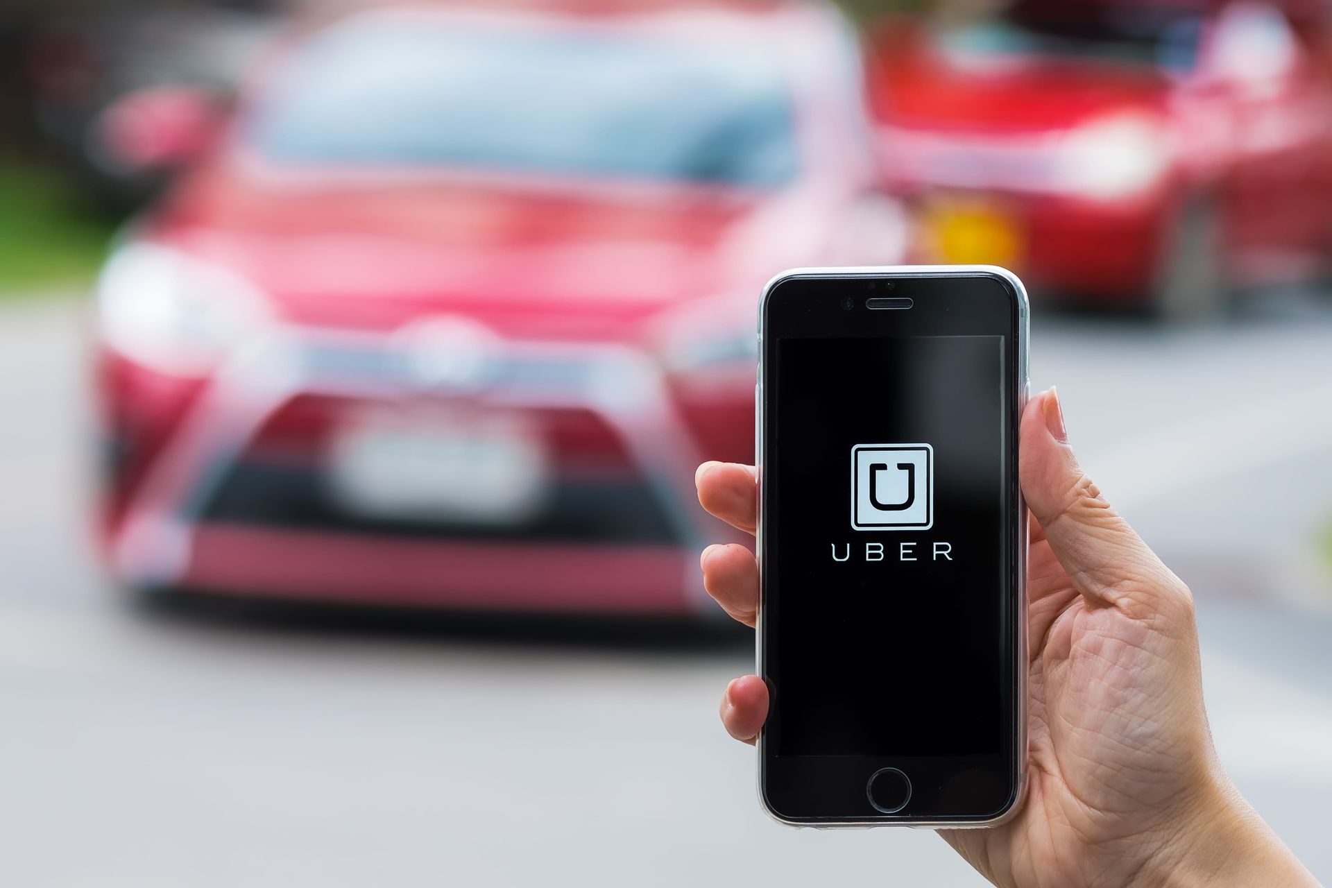 A hand holds a smartphone displaying the Uber app logo, with a blurry red car in the background.
