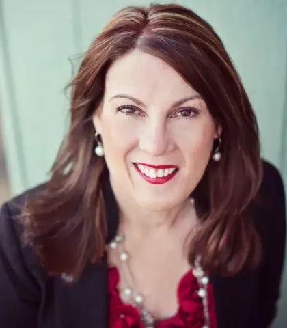 A woman wearing a black jacket and pearl earrings smiles for the camera