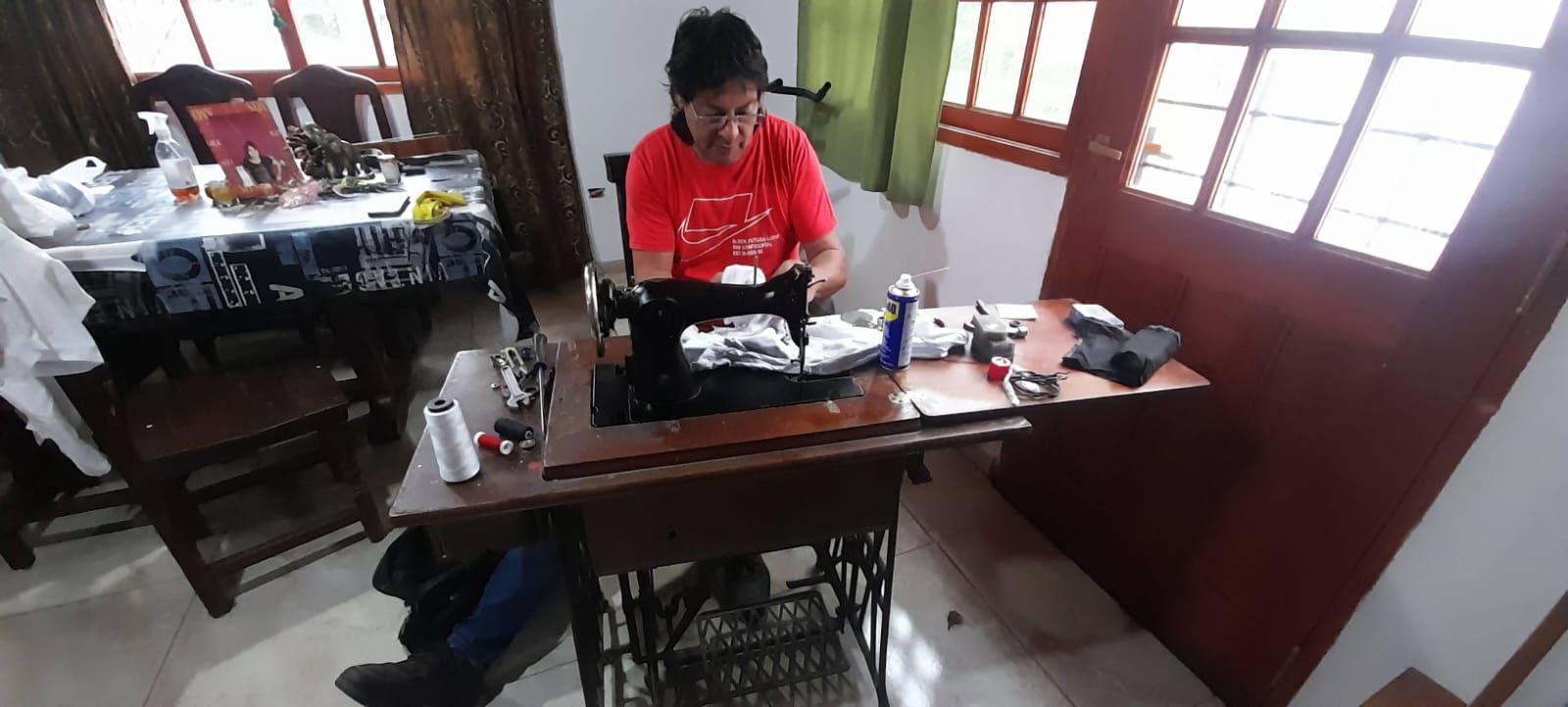 Persona con camisa roja trabajando en una máquina de coser en una habitación pequeña con tela y herramientas sobre una mesa.
