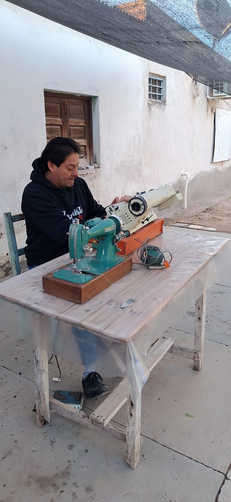 Mujer cosiendo en una máquina verde sobre una mesa al aire libre junto a una pared blanca.
