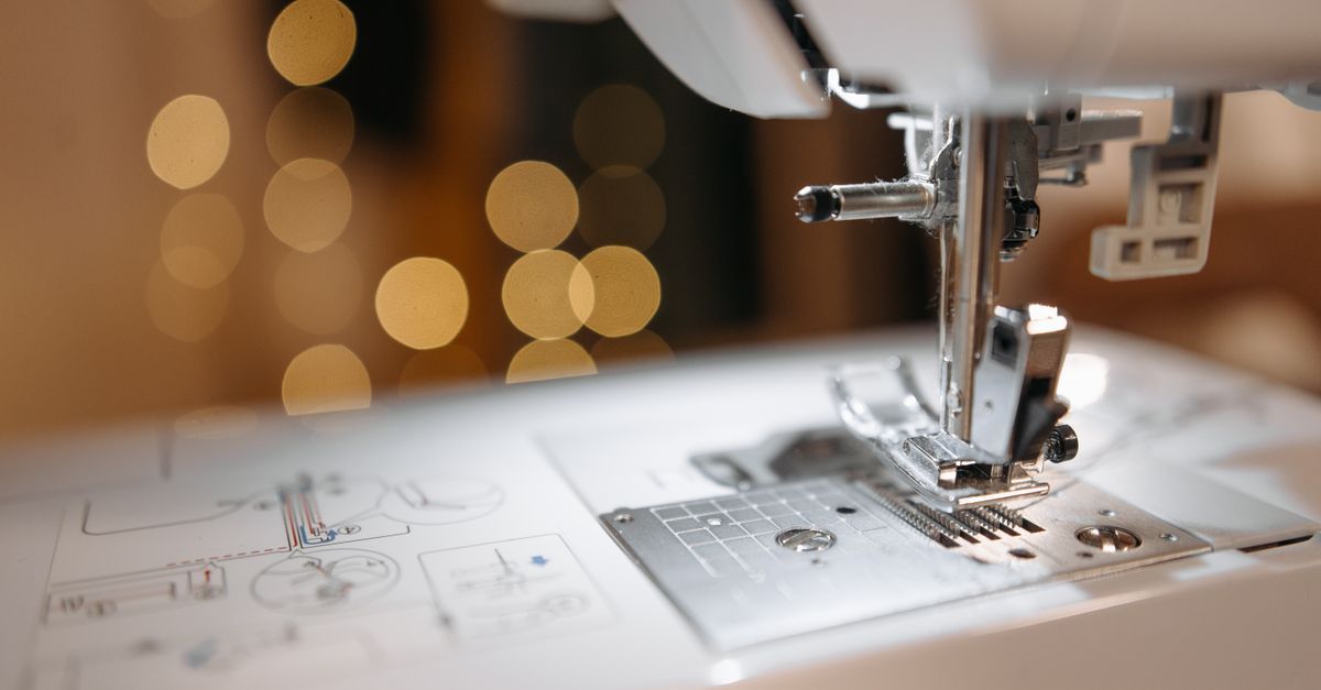 Primer plano de una máquina de coser cosiendo tela blanca con luces bokeh cálidas al fondo.